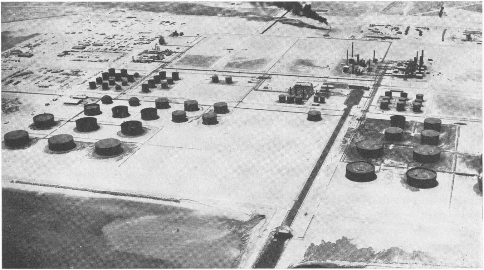 Foto antiga em preto e branco da refinaria de Ras Tanura, com tanques e chaminés à beira-mar